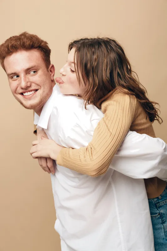 Young couple in casual clothes share a playful moment, woman hugging man from behind and pretending to kiss his cheek. Concept of love, friendship, happiness, and relationship.
