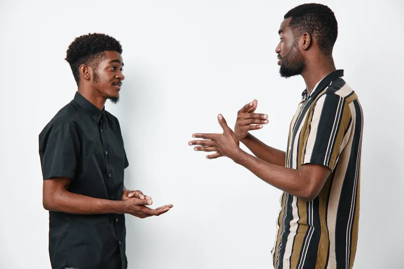 Two black men with dark skin having a conversation standing against white background. Casual clothing, communication, discussion, friendship, interaction, young adults, studio portrait.
