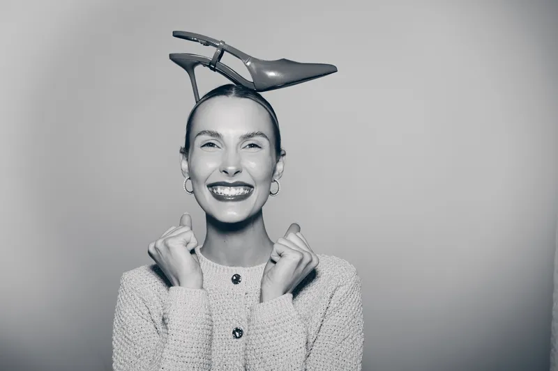 Smiling white woman with pale skin holds high heel shoe on head, wearing cozy sweater, studio portrait on light background. Concept of fun and fashion.