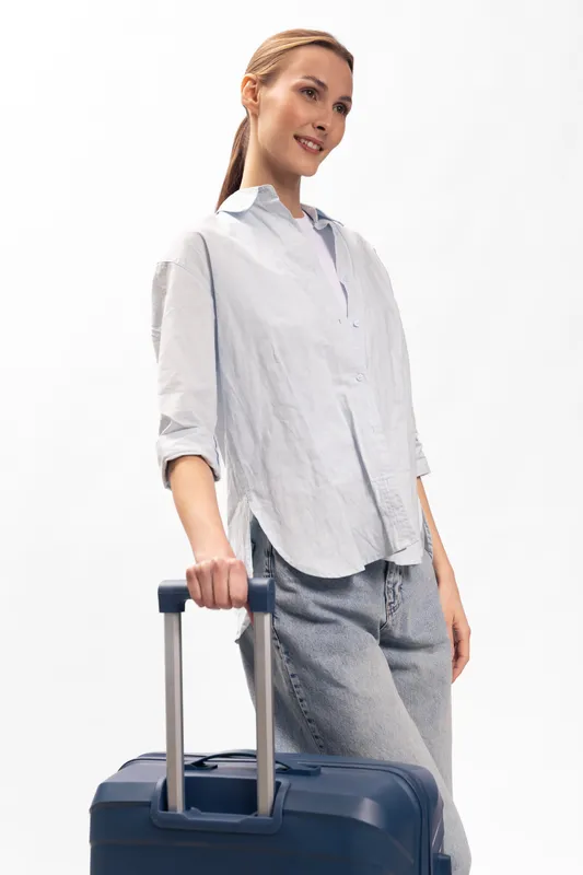 Young woman with suitcase stands smiling against white background. She wears casual clothes and holds handle of blue luggage. Concept of travel, vacation, and lifestyle.