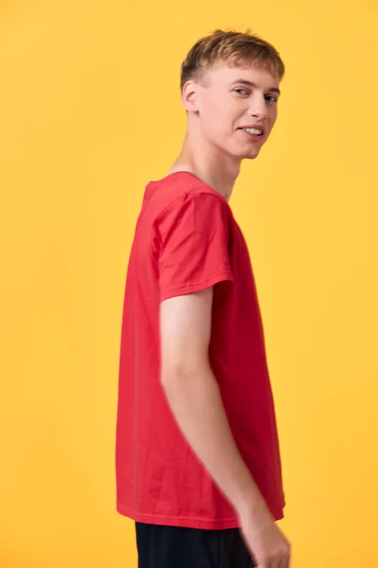 Young man in a red shirt turns slightly to the side against a bright yellow background, presenting casual fashion, confidence, and a friendly, approachable vibe in studio light.