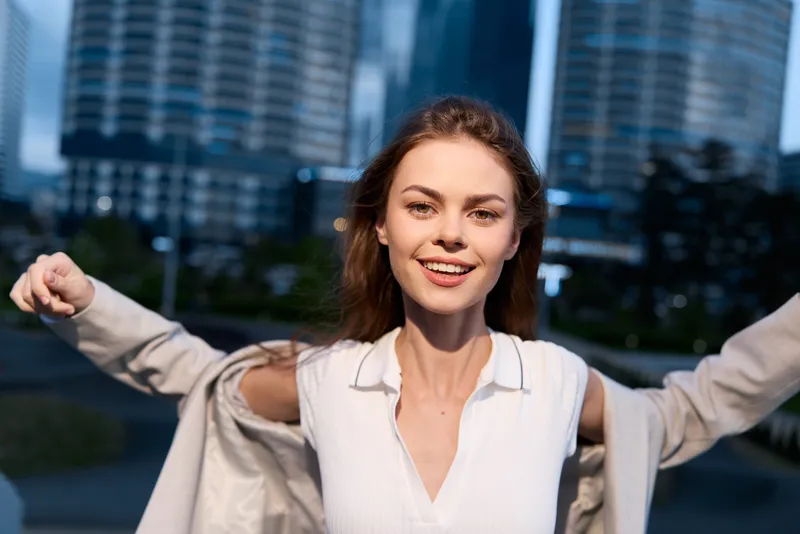 Young woman in white shirt smiling outdoors with open coat and city buildings in background at dusk, positive energy, casual style, urban fashion