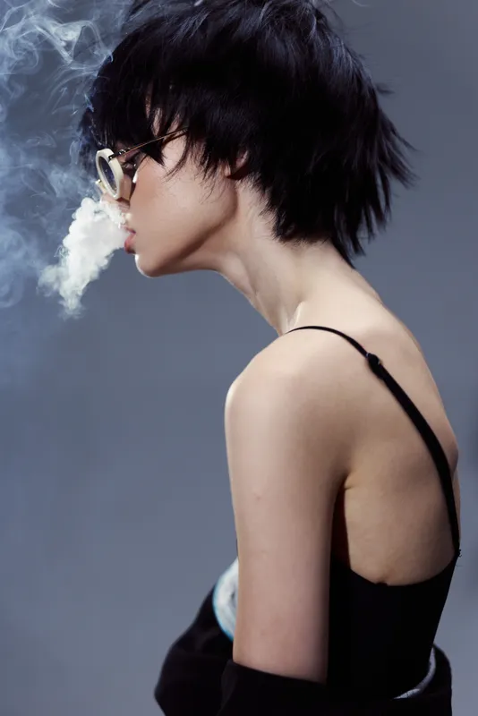 Young woman with short black hair wearing glasses exhales smoke against a gray background in a minimalistic style.