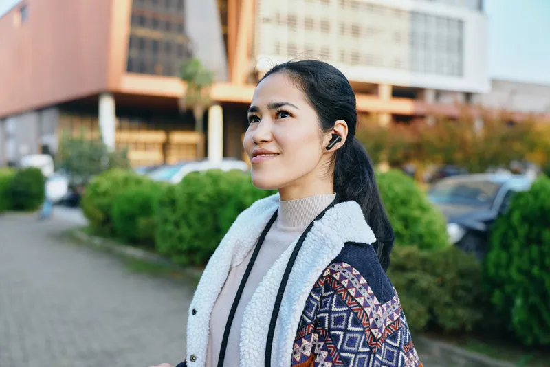 Woman wearing a patterned coat and light sweater stands outdoors on an urban street, listening with wireless headphones, looking up with a confident hopeful smile.
