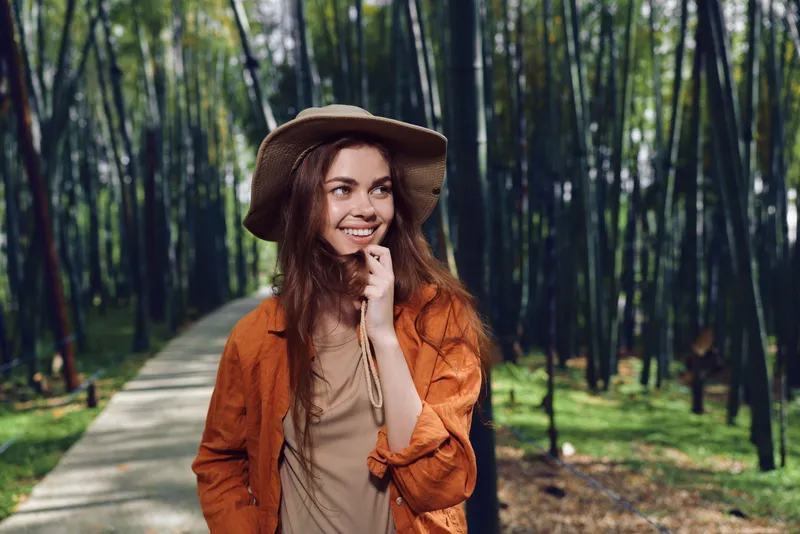 woman smile hat bamboo forest path walking nature portrait of a happy traveler in orange jacket on a scenic trail, candid outdoor portrait with sunlight, greenery and relaxed joyful expression