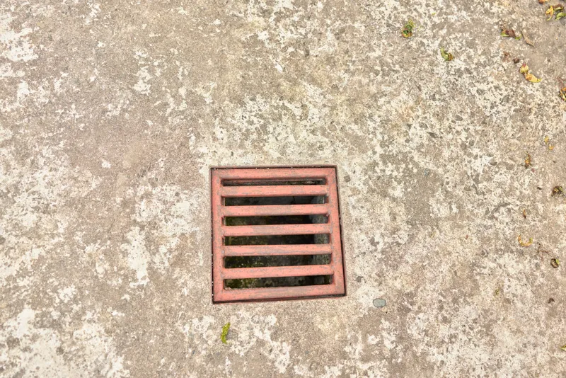 Close up of a square metal drain on rough concrete pavement, outdoor urban scene, neutral tones, weathered surface, subtle shadows, minimalist composition, everyday street detail, natural lighting