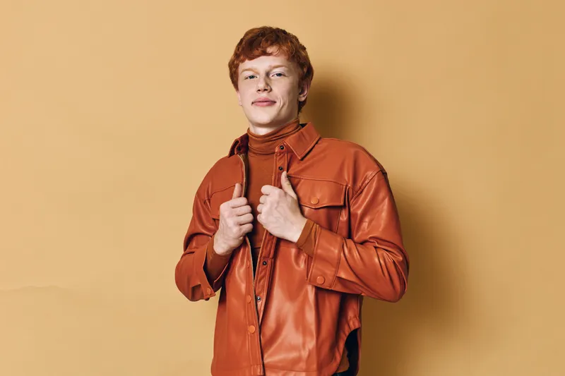 Young man with red hair wearing a brown leather jacket and turtleneck posing confidently with hands on collar against beige background in studio.