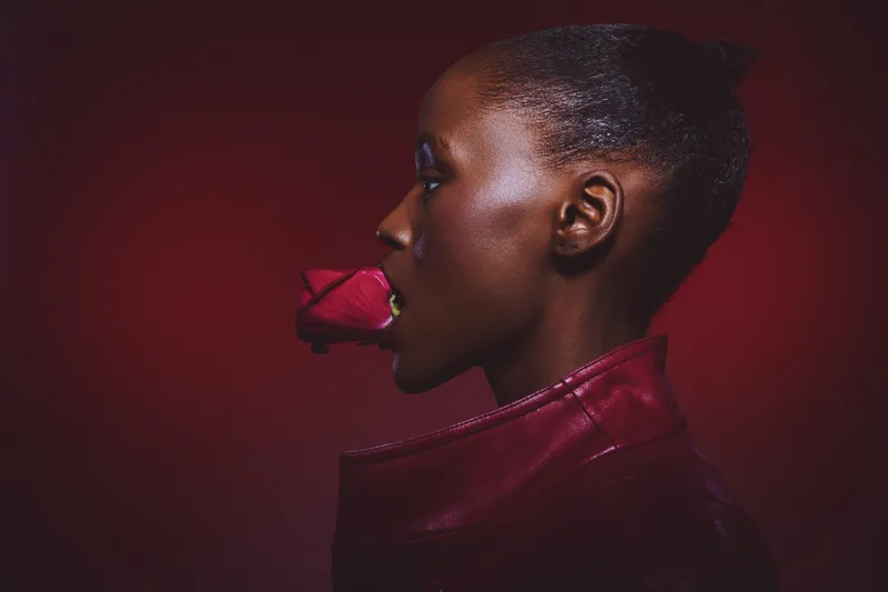 Profile of a dark skinned African woman holding a red rose in her mouth wearing a red leather jacket against a dark red background. Concept of beauty, elegance, and passion.