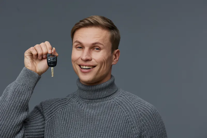 smiling caucasian man holding car key showing remote control in gray turtleneck sweater isolated on gray background cybersecurity concept