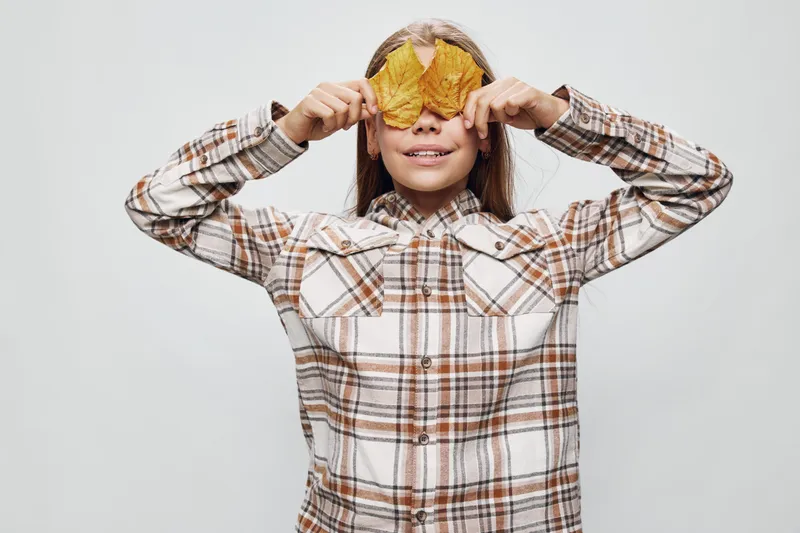 Child girl holding large yellow autumn leaves over eyes smiling with casual plaid shirt on light background for autumn season fun and nature play.