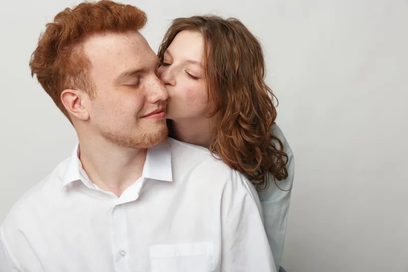 Young couple in casual clothes share a tender moment as woman kisses man on cheek. Intimate love and affection concept with soft natural light and neutral background.