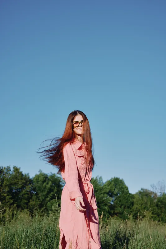 Young woman in a pink dress smiling joyfully outdoors with flowing hair, against a clear blue sky and green trees, representing freedom and happiness