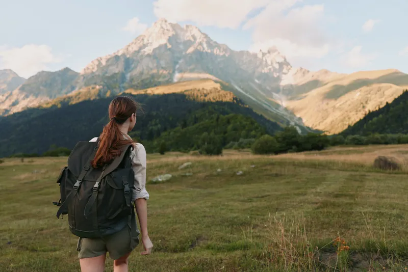 A young woman with a backpack stands on a grassy field looking at a mountain range under a partly cloudy sky. Concept of outdoor adventure, hiking, and lifestyle.
