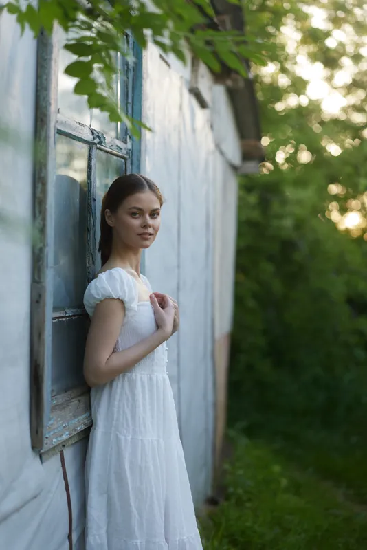 Young woman in a white dress standing by an old window, surrounded by greenery, exuding a serene and contemplative mood, ideal for nature inspired themes