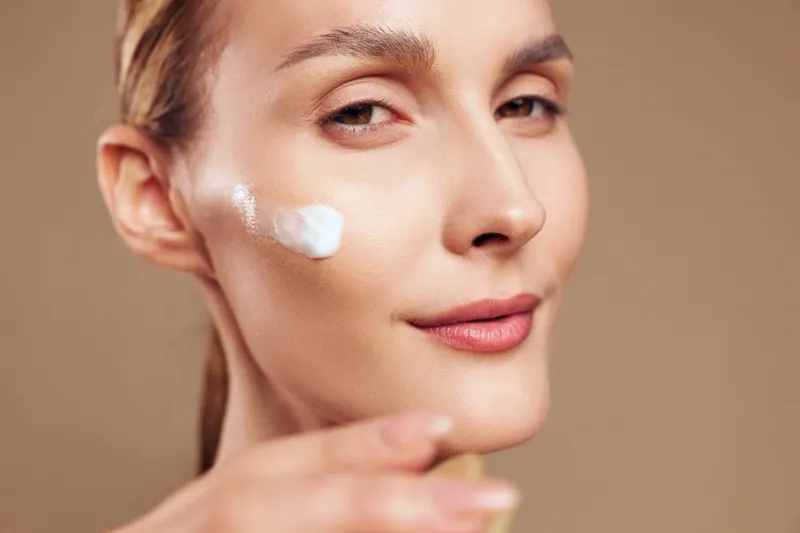 Woman in close up applying white face cream to cheek, calm soft smile, studio lighting on warm beige background, skincare wellness and self care concept, gentle skin texture focus
