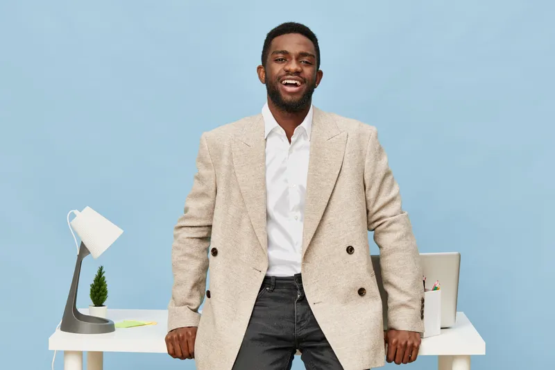 Smiling happy young African American man in beige blazer and white shirt standing in modern office with blue background. People lifestyle concept, professionalism, positive attitude, work environment,