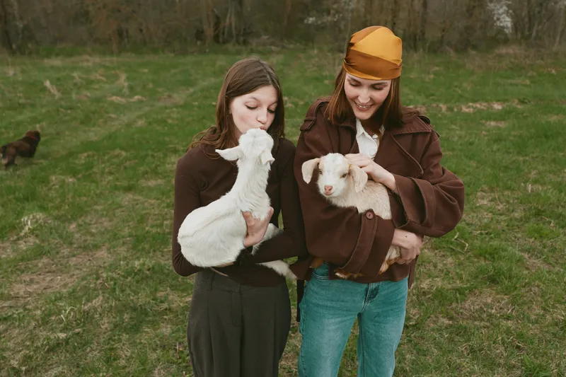 Two women outdoors hold white goat kids in a grassy field, sharing a gentle moment. A lifestyle scene highlighting companionship, animal care, and rustic farm life in daylight.