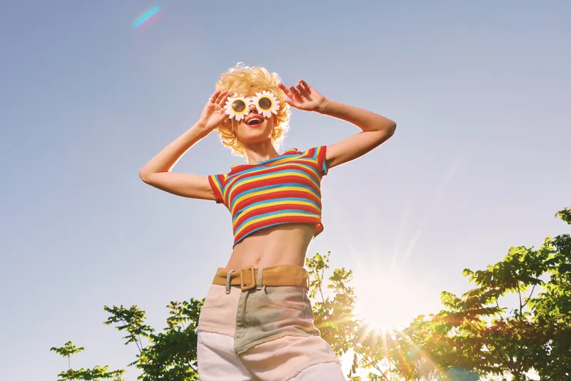 Young woman in striped croptop and high waist shorts smiles under bright sun, outdoors, capturing carefree summer lifestyle with retro film color and joyful vibe.