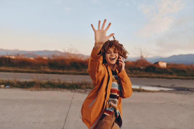 Woman lifestyle outdoors, laughing and smiling while waving, walking on a quiet road at sunset, warm golden light, soft breeze, casual jacket, scenic mountains background