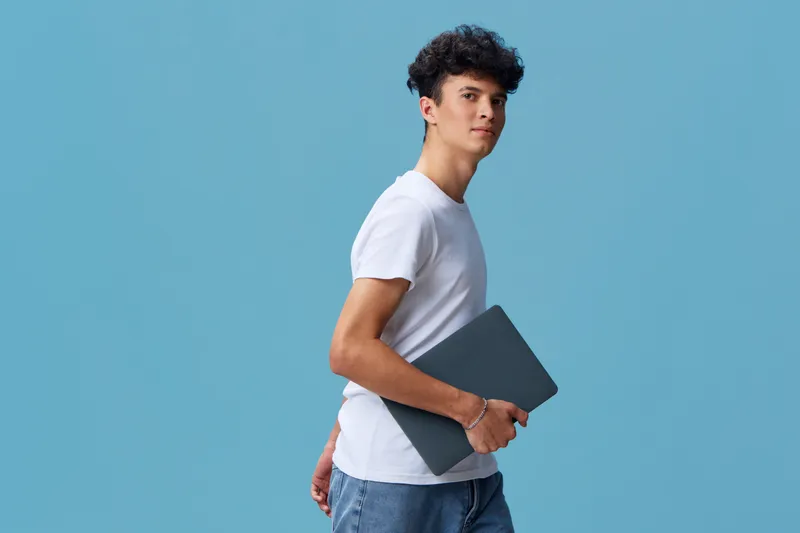 young man holding laptop, wearing casual white t-shirt and jeans, standing confidently against solid blue background