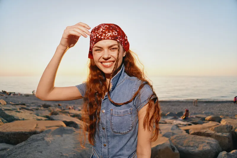 lifestyle woman in modern boho-western denim street style smiling with red bandana by rocky beach in warm film color lighting outdoors