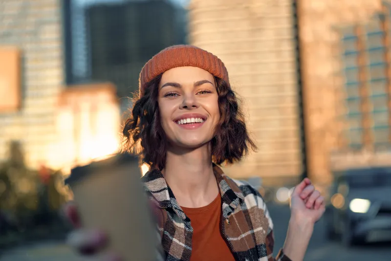 Woman portrait smiling with coffee cup in urban street at golden hour, candid beanie style and casual jacket, authenticity, mindful living, golden hour glow, emotional storytelling