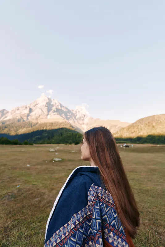 Woman back facing mountain landscape in a meadow, nature outdoors scene with patterned jacket. Traveler portrait with long hair, peaceful field and distant alpine horizon.