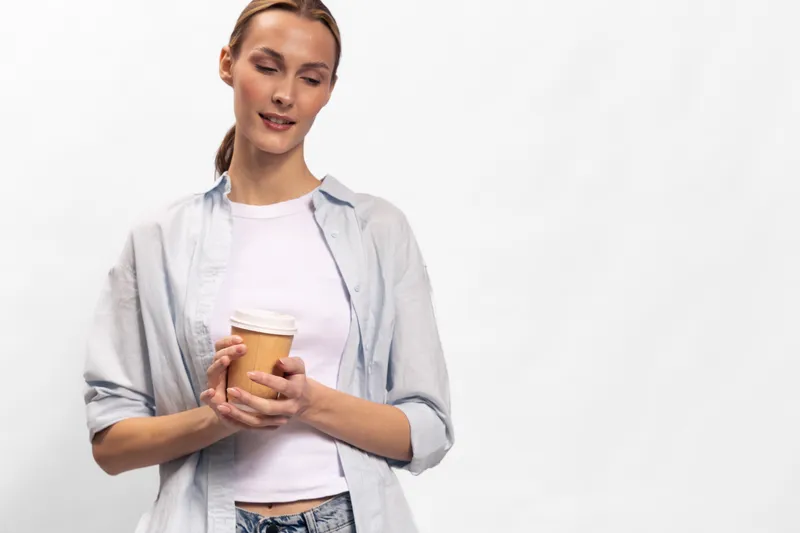 Young woman in casual clothes holds disposable coffee cup, looks down with soft smile. Concept of relaxation, break, and everyday lifestyle in minimalistic studio environment.