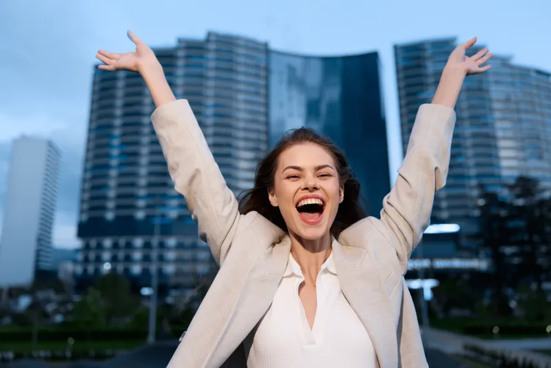 Happy businesswoman celebrating success in urban environment, wearing a stylish blazer and expressing joy with arms raised amidst modern buildings.