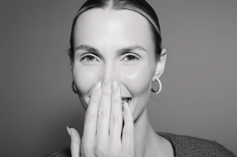 Smiling white woman with light skin covers mouth with hand in close-up portrait on grey background. Concept of happiness, surprise, and natural beauty in studio lighting.