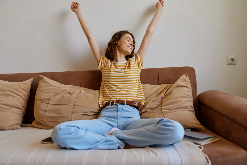 A joyful woman sits cross legged on a sofa in a cozy living room, arms raised in celebration. Bright natural light highlights a relaxed home lifestyle moment, suitable for branding and lifestyle