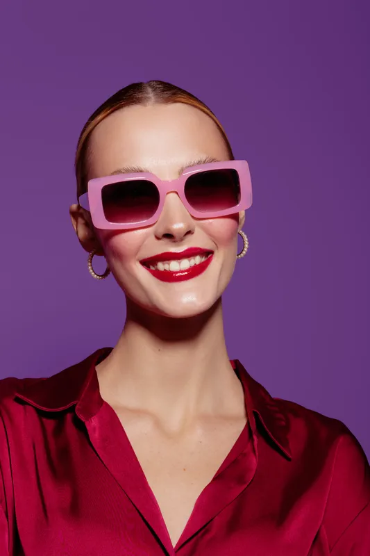 Smiling woman with pink sunglasses and red lipstick wears red satin blouse on purple background. Fashion portrait with bright makeup and trendy accessories for style concept.