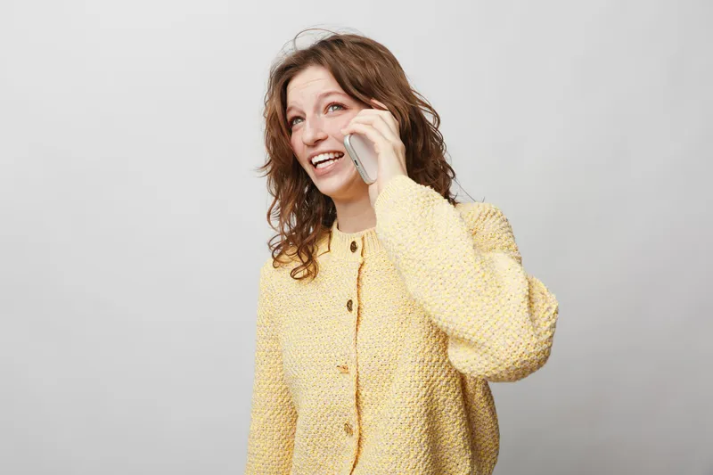 Smiling woman with curly hair wearing yellow sweater talks on smartphone. She looks happy and engaged, isolated on light grey background. Concept of communication, technology, and positive mood.