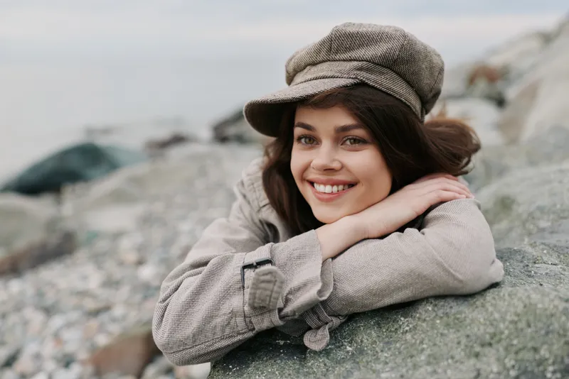 Smiling woman in beige plaid jacket and cap leans on rock outdoors. Natural light highlights her relaxed pose and joyful expression in a casual, autumn setting.