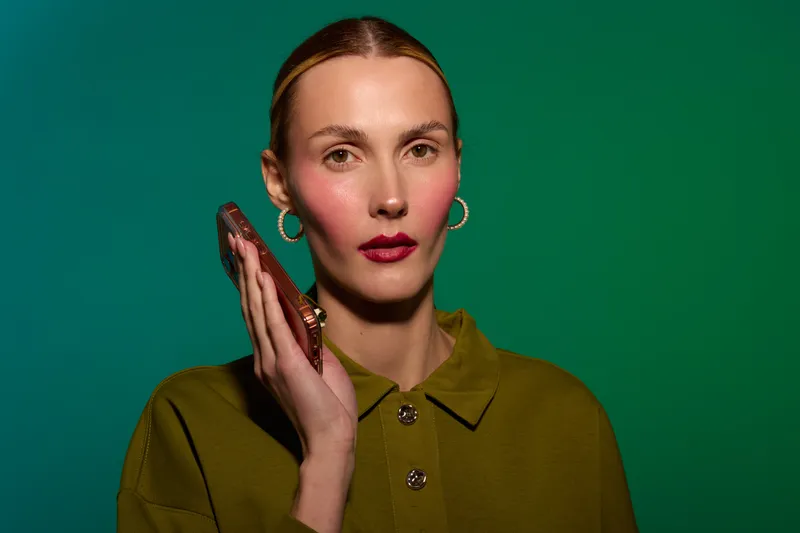 Young woman with red lipstick and hoop earrings holds a smartphone near her face against a green background. Concept of communication, technology, and modern lifestyle.