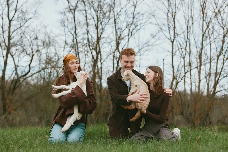 Three friends outdoors with playful dogs in a grassy park, warm casual outfits, laughter and gentle pet affection, natural daylight, relaxed lifestyle moment capturing friendship and animal