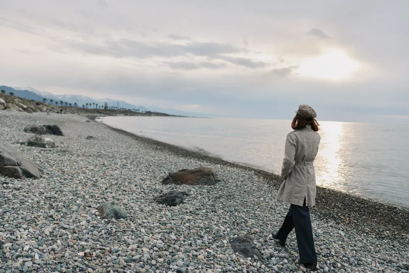 Woman in coat walks alone on rocky beach during overcast sunset. Calm sea and cloudy sky create peaceful mood. Concept of solitude, reflection, and nature outdoors.