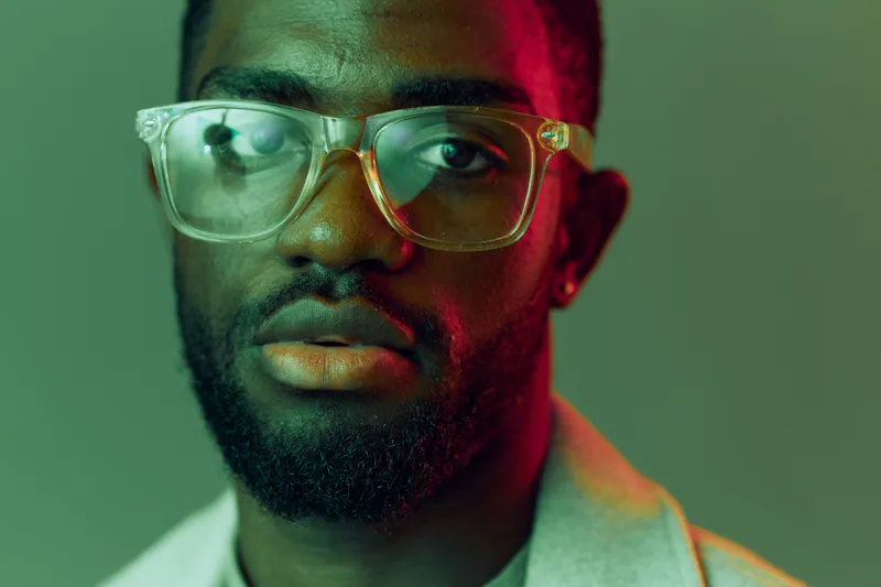 Portrait of a young African man wearing trendy glasses with an expressive gaze, set against a vibrant green and pink background, emphasizing modern fashion