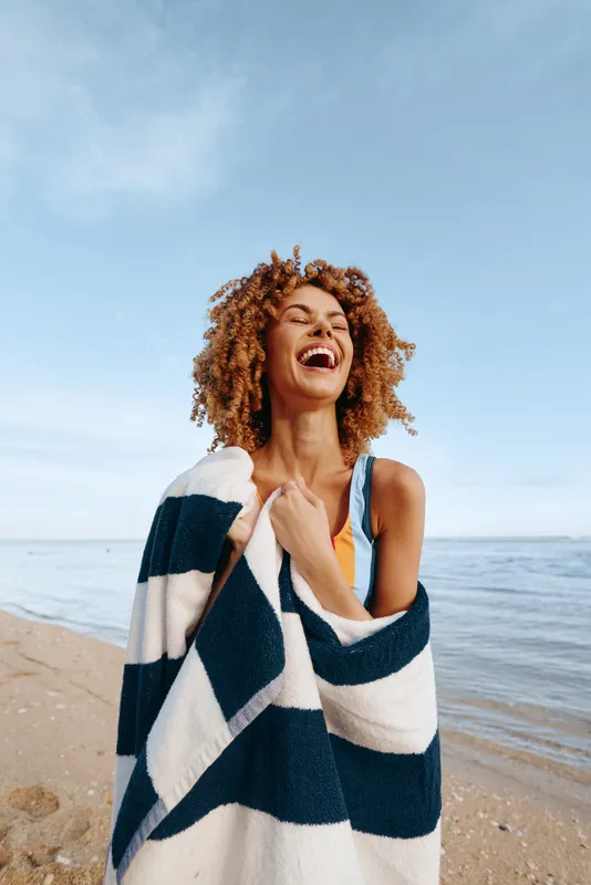 Woman curly hair beach laughing, wrapped in striped towel, smiling in bright sunlight, ocean horizon behind, calm waves nearby, summer vacation outdoor portrait, fresh air, carefree joy
