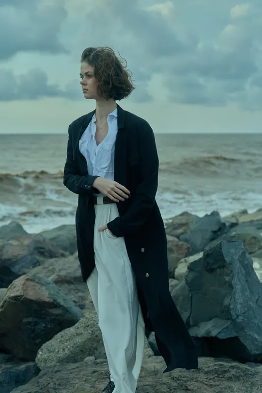 Woman in a long black coat over white trousers stands on a rocky shoreline, wind lifting hair, creating a dramatic seascape with fashion forward style.