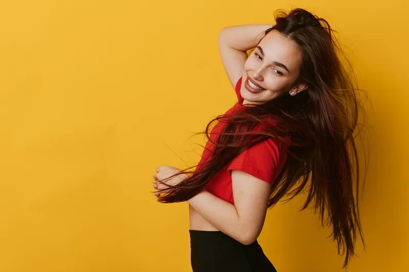 Young woman with long hair smiling and posing against yellow background wearing red shirt and black pants. Happy female model with natural makeup and carefree expression in studio.