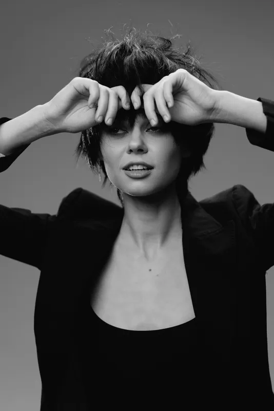 Black and white portrait of a young woman with short hair adjusting her bangs, wearing a dark blazer, expressing a relaxed mood.