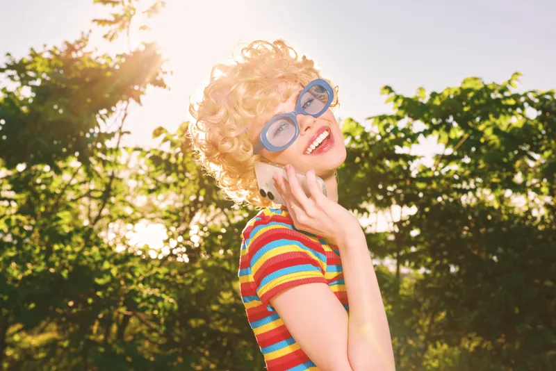 Joyful young woman in a colorful striped shirt and oversized sunglasses smiles under sunlit trees, enjoying a carefree outdoor moment with warm breeze
