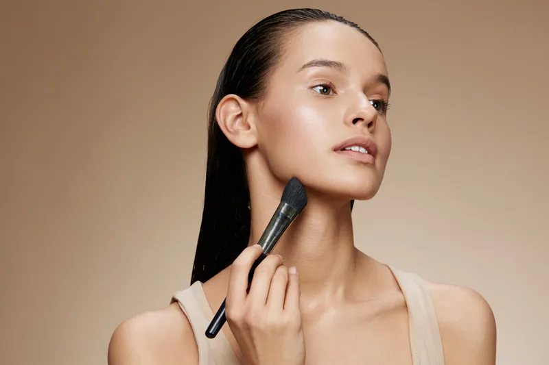 A young woman applying makeup with a brush, showcasing radiant skin and a natural look, perfect for beauty and wellness concepts in photography