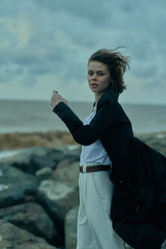 Woman standing on a rocky coast, jacket blowing in the wind, bag over shoulder, looking toward the sea under a moody sky by the sea breeze