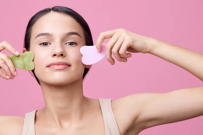 Young woman with natural beauty holding facial massage tools, promoting skincare wellness and relaxation, in a bright pink background for a fresh aesthetic