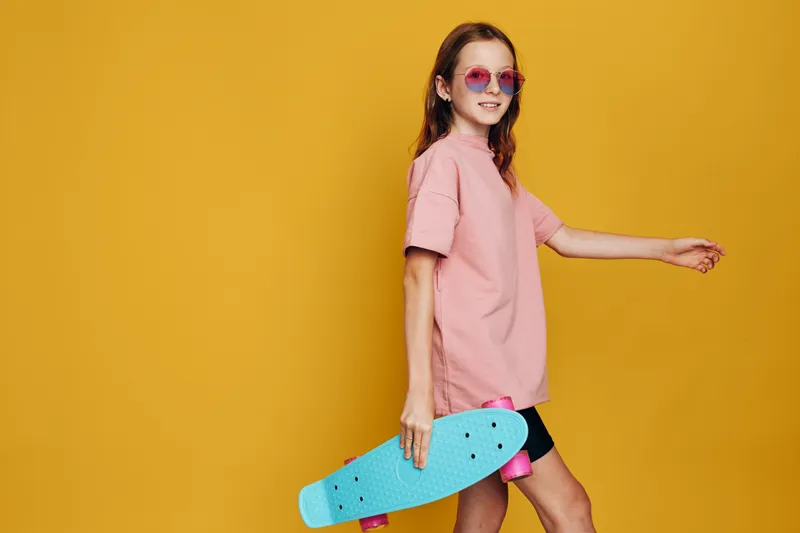 Young girl in pink t-shirt holds blue skateboard and walks against yellow background. Casual style with sunglasses. Concept of youth and leisure.