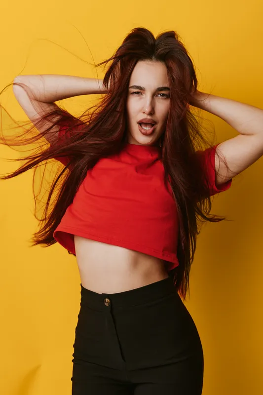 Young woman with long hair wearing red crop top and black pants posing with hands behind head on yellow background for fashion and style portrait.