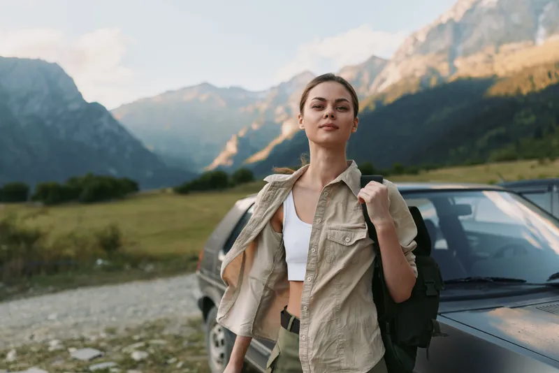 Young woman lifestyle traveler standing near car in mountain landscape confident expression casual outfit natural light outdoor adventure