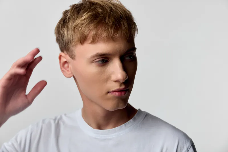 Young man with light hair in a white tee gazes to the side, hand raised near the head, studio lighting and a calm expression creating a casual yet thoughtful mood for fashion or lifestyle projects.